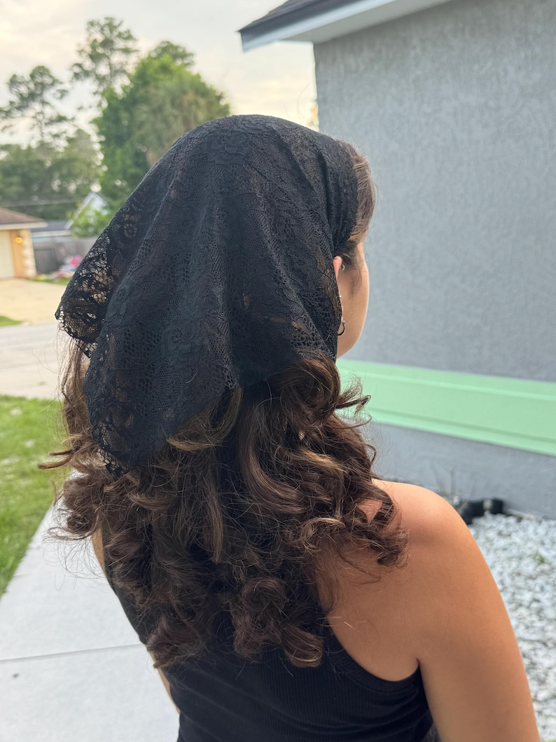 May include: A black triangular headscarf made of delicate lace fabric with a floral pattern. The headscarf is tied at the back of the head, covering the top portion of the hair, and features scalloped edges along the perimeter of the fabric.