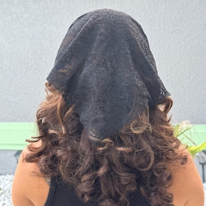 May include: A black triangular head covering made of delicate floral lace fabric. The accessory features a detailed, intricate pattern throughout the material and is worn draped over the top of the head, tapering to a point at the back.