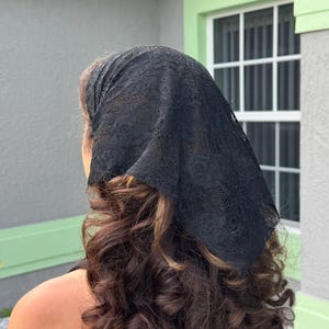 May include: A black triangular headscarf made of delicate lace fabric with a floral pattern. The headscarf is tied securely at the back of the head, covering the crown and draping down towards the nape of the neck over wavy brown hair.