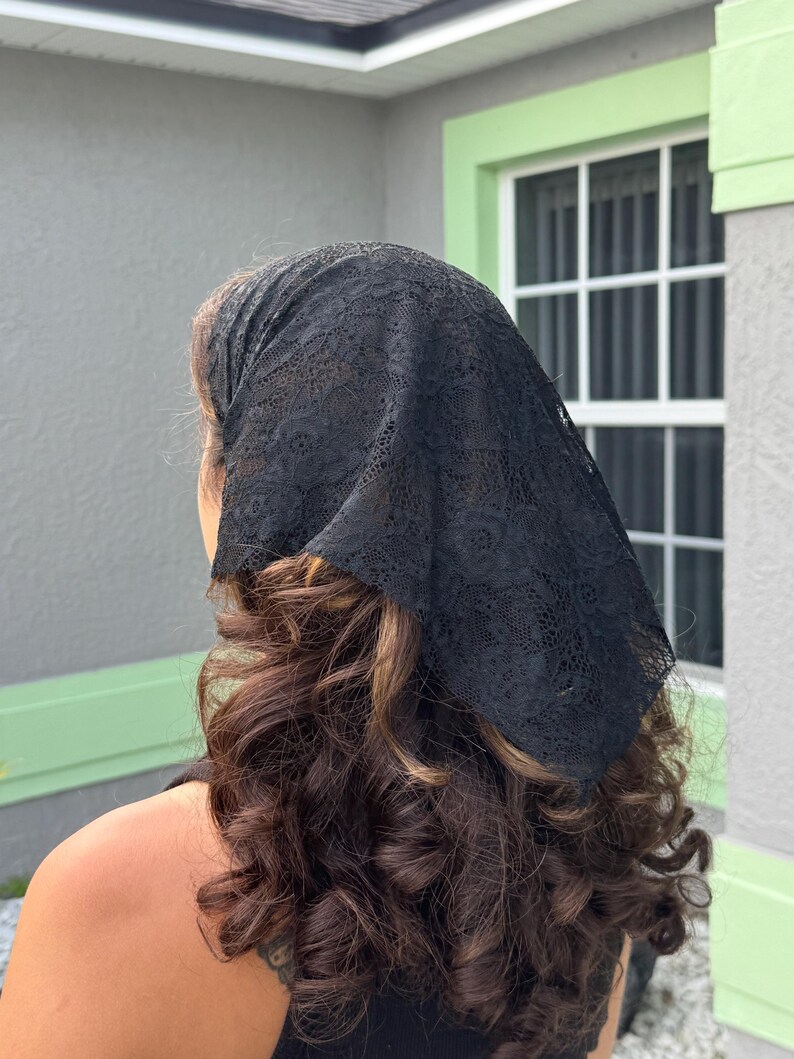May include: A black triangular headscarf made of delicate lace fabric with a floral pattern. The headscarf is tied securely at the back of the head, covering the crown and draping down towards the nape of the neck over wavy brown hair.
