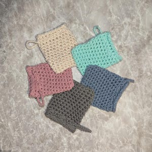 May include: A collection of six handmade crochet washcloths in various colors: cream, light blue, pink, gray, and navy blue. Each washcloth has a loop for hanging. The washcloths are arranged in a circular pattern on a marble surface.