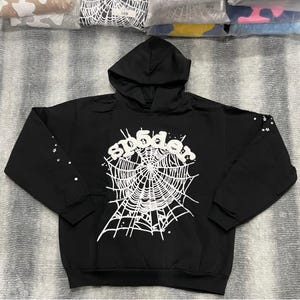 Könnte beinhalten: Schwarzer Kapuzenpullover mit weißem Spinnennetz-Design und dem Wort "spider". Die Ärmel sind mit kleinen weißen Sternen verziert. Ein lässiges, bequemes Oberteil.