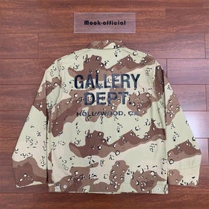 Puede incluir: Chaqueta de camuflaje marr&oacute;n y beige con el texto "GALLERY DEPT" y "HOLLYWOOD, CA" impreso en la espalda. La chaqueta tiene una botonadura frontal y un cuello. La marca "Mook-official" est&aacute; impresa en una peque&ntilde;a etiqueta rectangular.