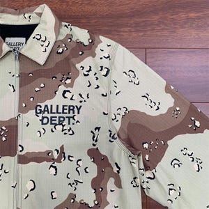 Puede incluir: Chaqueta de camuflaje en tonos beige, marr&oacute;n y negro, con cierre de cremallera. La chaqueta muestra el texto "GALLERY DEPT." en negro. Tiene cuello y mangas largas.