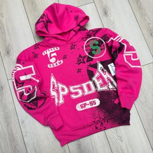 May include: A bright pink hooded sweatshirt with the word "SPDER" in large white letters across the front. The design includes black star graphics, the number "5", and the text "SPDER TEAM". The sweatshirt also features the text "SP-55".