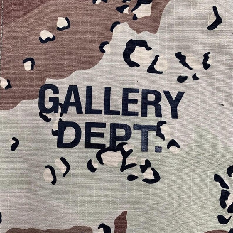 Puede incluir: Primer plano de una tela con un estampado de camuflaje en tonos marr&oacute;n, beige y negro. Las palabras "GALLERY DEPT." est&aacute;n impresas en letras negras grandes y en negrita en la tela. La tela tiene una apariencia texturizada.