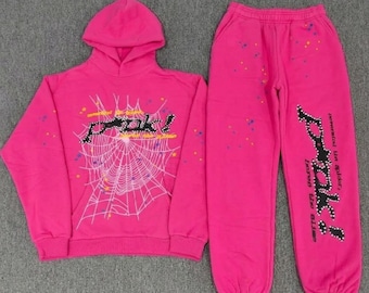 May include: A bright pink hooded sweatshirt and matching sweatpants set. The hoodie features a white spiderweb graphic and the text "peak!" in black with white dots. The pants have the same "peak!" text and scattered yellow and blue paint splatters.