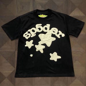 Könnte beinhalten: Schwarzes T-Shirt mit dem Wort "sp5der" in cremefarbener Schrift im Blasenstil und cremefarbenen Sternen. Das T-Shirt hat einen Rundhalsausschnitt und kurze Ärmel. Das T-Shirt ist aus weichem Material.