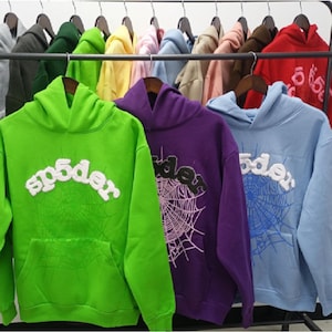 May include: A rack of hooded sweatshirts in various colors, including green, purple, and blue. Each hoodie features a spider web design and the word "sp5der" in a stylized font. The hoodies have a front pocket and a drawstring hood.