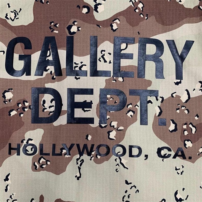 Puede incluir: Primer plano de una tela con un estampado de camuflaje en tonos marr&oacute;n, beige y crema. Las palabras "GALLERY DEPT." est&aacute;n impresas en letras grandes y oscuras, con "HOLLYWOOD, CA." debajo.