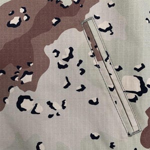 Puede incluir: Primer plano de una secci&oacute;n de tela de camuflaje. El patr&oacute;n presenta formas irregulares en beige, marr&oacute;n y negro sobre un fondo gris claro. Un bolsillo rectangular con cremallera beige es visible en el lado derecho.