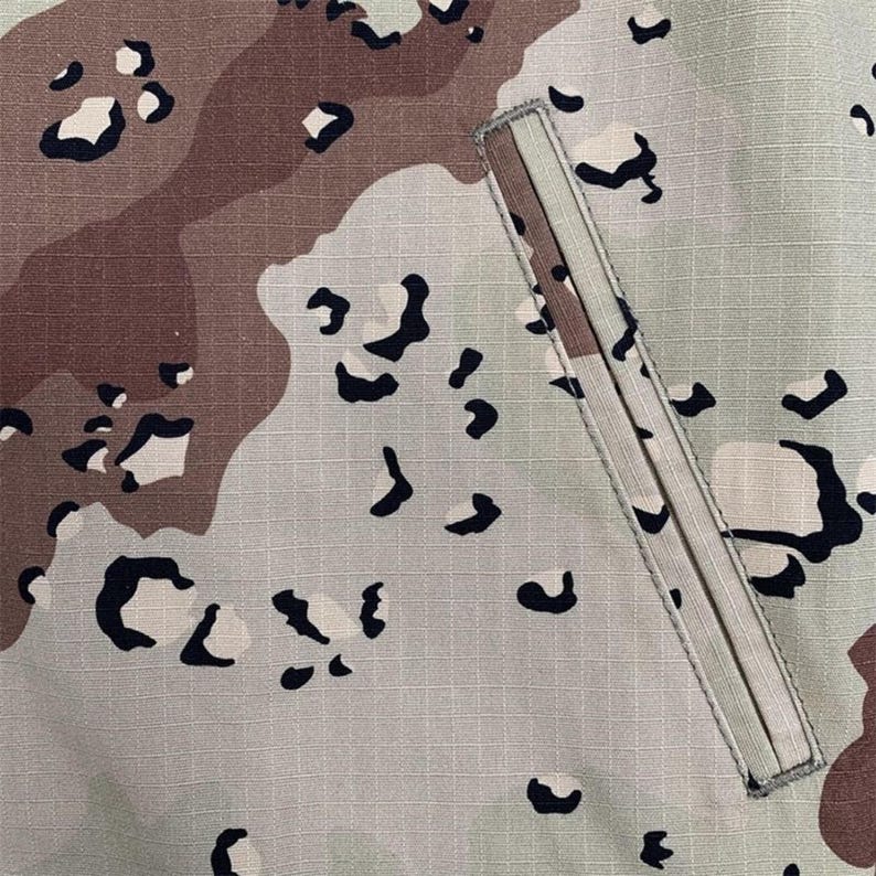 Puede incluir: Primer plano de una secci&oacute;n de tela de camuflaje. El patr&oacute;n presenta formas irregulares en beige, marr&oacute;n y negro sobre un fondo gris claro. Un bolsillo rectangular con cremallera beige es visible en el lado derecho.