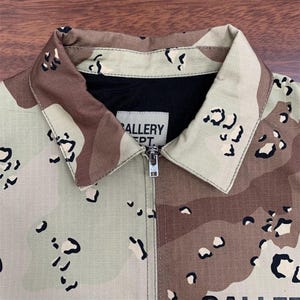 Puede incluir: Primer plano de una chaqueta de camuflaje con cierre de cremallera. La chaqueta presenta un patr&oacute;n de camuflaje des&eacute;rtico en tonos beige, marr&oacute;n y negro. El cuello y la parte superior de la chaqueta son visibles, con el texto "GALLERY DEPT" en una etiqueta.