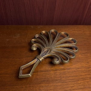 May include: A brass-toned metal trivet featuring a fan-shaped design with curved, open-work radiating spokes and a diamond-shaped handle. The object has a weathered, aged finish with visible dark patina in the recessed areas of the metal.