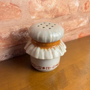 May include: A ceramic salt shaker with a mushroom-shaped design. The top is light gray with several holes, and the middle section has a ruffled edge. The bottom portion is white with a floral pattern and red accents. The shaker sits on a wooden surface.