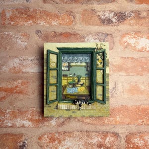 May include: A framed, three-dimensional artwork depicting a kitchen scene viewed through an open window. The window frame is green, and a black and white cat rests on the windowsill. The kitchen features a table with a blue and white checkered tablecloth.