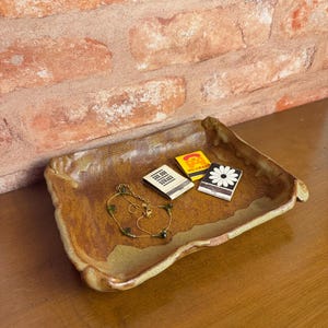 May include: A rectangular ceramic tray with a mottled brown and tan glaze featuring uneven, raised edges. Inside the tray rests a delicate gold-toned chain necklace with small green beads, a yellow matchbox, and a black matchbox with a white daisy.