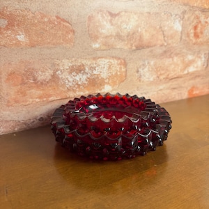 May include: A circular, deep red glass ashtray featuring a textured hobnail pattern across its exterior surface. The piece has a scalloped, notched rim design and a translucent finish that allows light to pass through the vibrant red material.