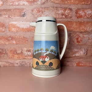 May include: Cream-colored vintage thermos with a landscape scene featuring a house, trees, and a blue sky. The thermos has a silver rim and handle. It is a classic design for keeping beverages hot or cold.