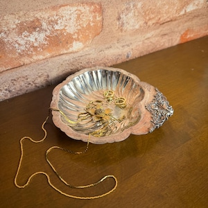May include: A silver-colored, shell-shaped trinket dish with a decorative edge. The dish contains several gold rings and a gold chain necklace. The dish sits on a dark wooden surface, with a brick wall in the background.