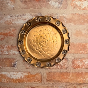 May include: A round, gold-colored metal tray with a floral design in the center and a scalloped edge. The edge is decorated with small, embossed flower shapes. The tray is displayed against a brick wall.
