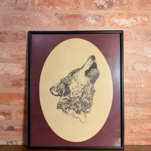 May include: A framed wall art piece featuring a black ink line drawing of a wolf howling. The drawing is set against a cream-colored oval background, which is surrounded by a deep maroon matting and enclosed within a simple black rectangular frame.