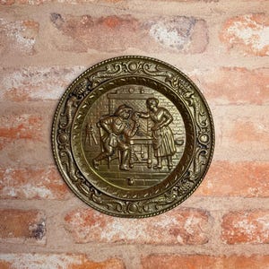 May include: A circular brass wall plate featuring a relief scene of two figures in a kitchen setting. The border is decorated with an embossed pattern of scrolls and circles. The central image depicts a person seated at a table while another person stands nearby, with a brick-patterned wall in the background.