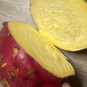 May include: A halved sweet potato with a vibrant red-purple skin and a bright yellow interior. The cut reveals the starchy texture of the vegetable. The sweet potato is on a wooden surface.