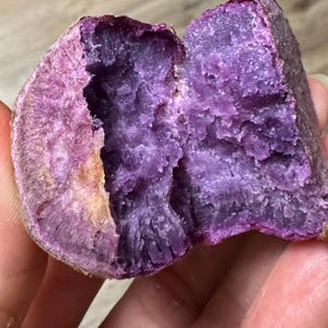 May include: A halved purple sweet potato showing a vibrant, deep violet interior flesh with a textured, starchy surface. The outer skin is a light brown, earthy color, contrasting with the intense purple color of the vegetable's inside.