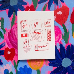 Könnte beinhalten: Eine weiße Karte mit roten Illustrationen von Streichholzschachteln, Streichhölzern und Text, einschließlich der Wörter "fm", "so", "glad", "we" und "matched". Die Karte befindet sich vor einem lebendigen floralen Hintergrund mit rosa, blauen und gelben Blumen.