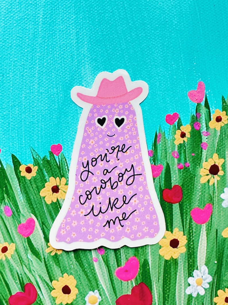 Puede incluir: Una pegatina caprichosa que presenta un fantasma con un sombrero de vaquero rosa. El fantasma est&aacute; cubierto de peque&ntilde;os estampados florales y tiene ojos en forma de coraz&oacute;n. El texto "you're a cowboy like me" est&aacute; escrito en el fantasma. El fondo incluye flores y hierba verde.