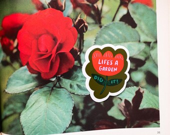 May include: A sticker with the text "Life's a Garden, Dig it!" in white and green, featuring a red flower design. The sticker is placed on a page with a vibrant red rose and green leaves.