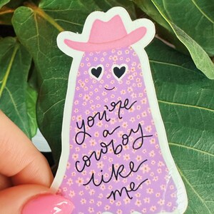 Puede incluir: Una pegatina con un fantasma que lleva un sombrero de vaquero rosa. El fantasma es morado con un estampado floral y ojos en forma de coraz&oacute;n. El texto "you're a cowboy like me" est&aacute; escrito en negro.