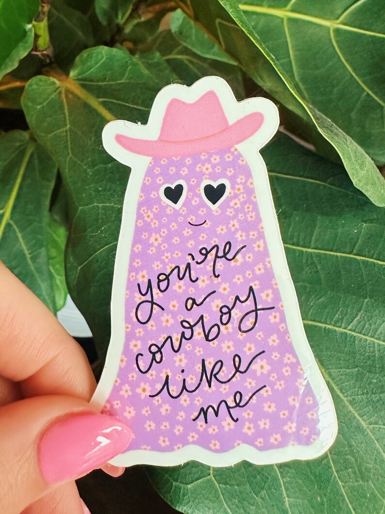 Puede incluir: Una pegatina con un fantasma que lleva un sombrero de vaquero rosa. El fantasma es morado con un estampado floral y ojos en forma de coraz&oacute;n. El texto "you're a cowboy like me" est&aacute; escrito en negro.