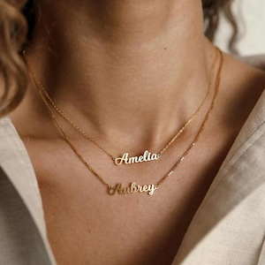 Pode incluir: São exibidos dois colares dourados com nomes, um com o nome "Amelia" e o outro "Aubrey". Os colares são em camadas, com correntes delicadas e letras cursivas. O fundo é o pescoço e a parte superior do peito de uma pessoa.