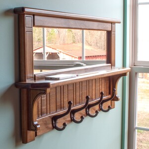 May include: A wooden wall-mounted shelf with a mirror and hooks. The shelf is stained a dark brown and has a decorative trim. The mirror is rectangular and reflects a view of trees and a red roof. There are five hooks on the bottom of the shelf.