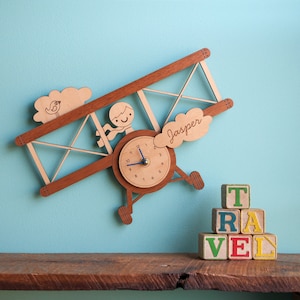 May include: Wooden airplane wall clock with a clock face and a pilot figure. The clock is painted brown and has a blue hand. The airplane has a cloud with the name "Jasper" written on it. The clock is displayed on a wooden shelf with a stack of colorful wooden blocks that spell "TRAVEL".