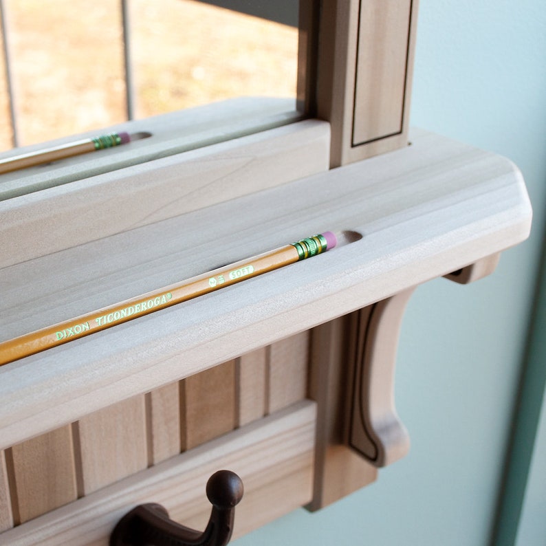 May include: A wooden wall-mounted shelf with a mirror, a hook, and a pencil holder. The shelf is made of light wood and has a natural finish. The pencil holder is a small groove in the shelf, and a yellow pencil is resting in the groove. The mirror is rectangular and has a simple frame.