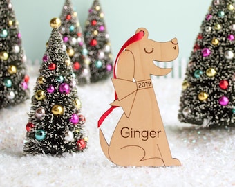 May include: A wooden dog ornament with a red ribbon and the name "Ginger" engraved on it. The ornament is dated "2019".