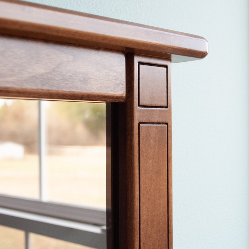 May include: Close-up of the top corner of a wooden mirror frame. The frame is a rich brown color and has a simple, elegant design.