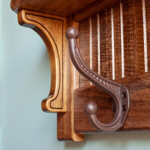 May include: A wooden coat rack with a brown finish and a single metal hook. The hook is made of dark brown metal and has a decorative design.