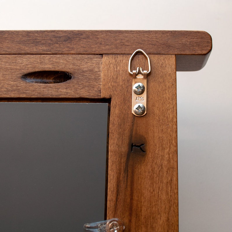 May include: A close-up of a wooden picture frame with a metal hanging hook attached to the back. The hook has two screws and the text "AMS" is visible on the hook.