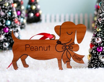 May include: A wooden dachshund ornament with the name "Peanut" and the year "2020" engraved on it. The ornament is brown and has a red ribbon around its neck.