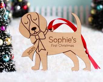 May include: A wooden Christmas ornament shaped like a beagle dog with a red ribbon around its neck. The ornament is personalized with the text "Sophie's First Christmas" and the year "2020".