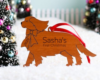 May include: A wooden dog ornament with the text "Sasha's First Christmas" and the year "2020" on it. The ornament is brown and has a red ribbon attached to it.
