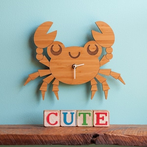 May include: A wooden crab clock with a white face and black hands. The clock is hanging on a light blue wall and has the word "CUTE" spelled out in colorful blocks below it.