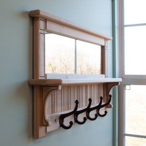 May include: A wooden shelf with a mirror and five hooks. The shelf is stained a light wood color and has a decorative trim around the mirror. The hooks are black metal and are attached to the bottom of the shelf.