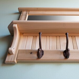 May include: A wooden wall-mounted shelf with a mirror and two hooks. The shelf is made of light wood and has a natural finish. The hooks are made of dark metal and are attached to the bottom of the shelf.