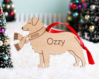 May include: A wooden Christmas ornament shaped like a dog wearing a scarf. The ornament is painted brown and has the text "2020" and "OzZy" engraved on it.