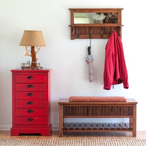 May include: A red wooden chest of drawers with six drawers and black handles. A wooden bench with a red cushion and a shelf underneath. A wooden wall mounted mirror with hooks and a red jacket hanging on one of the hooks. A lamp with a brown shade and a wooden base shaped like a tree.